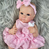 A pink reborn baby doll with painted hair and a bow, wearing a matching pink dress, lying on gray fur.