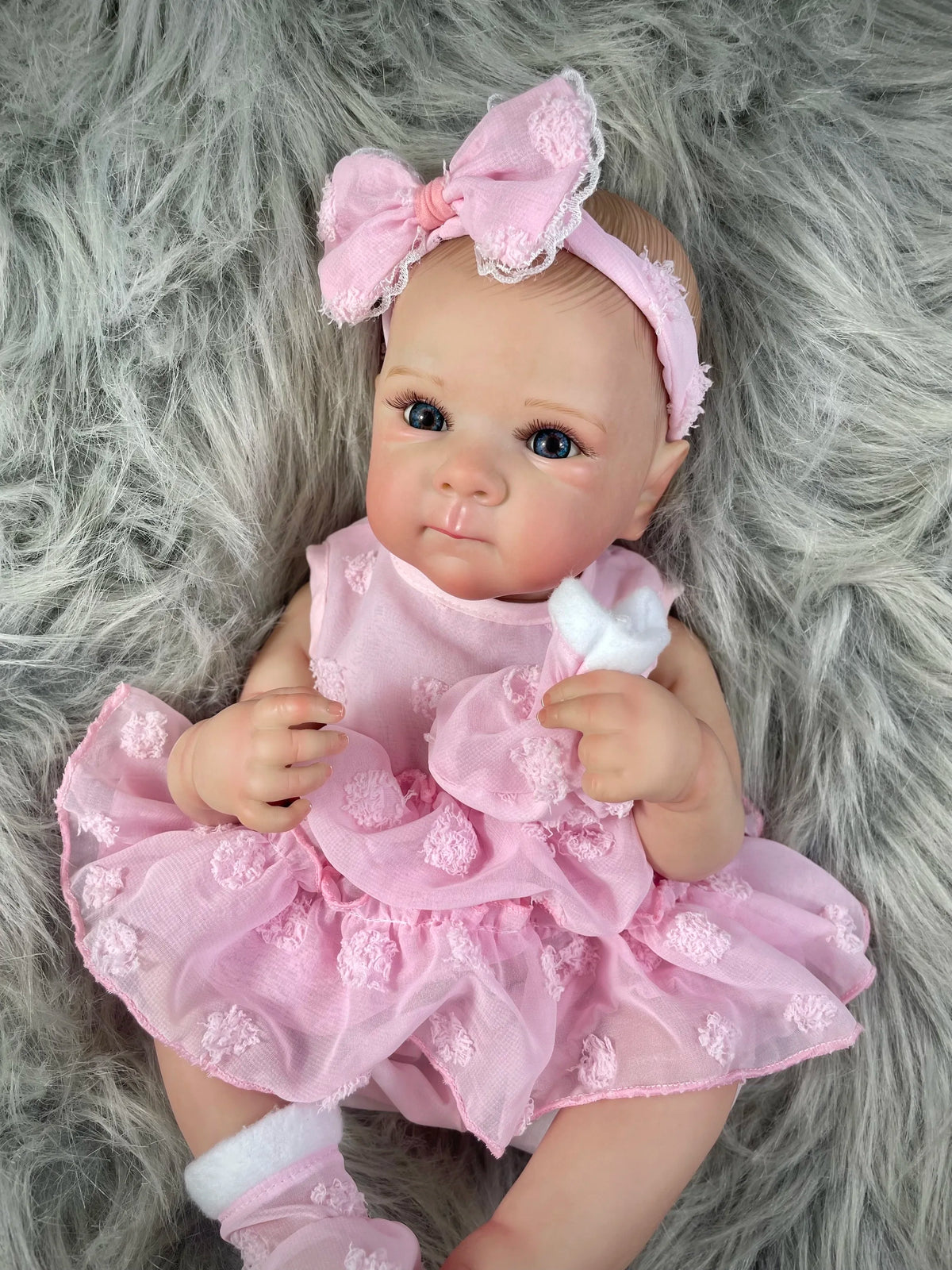A pink reborn baby doll with painted hair and a bow, wearing a matching pink dress, lying on gray fur.
