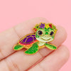A cute green turtle enamel pin featuring vibrant purple and yellow accents, held in a hand.