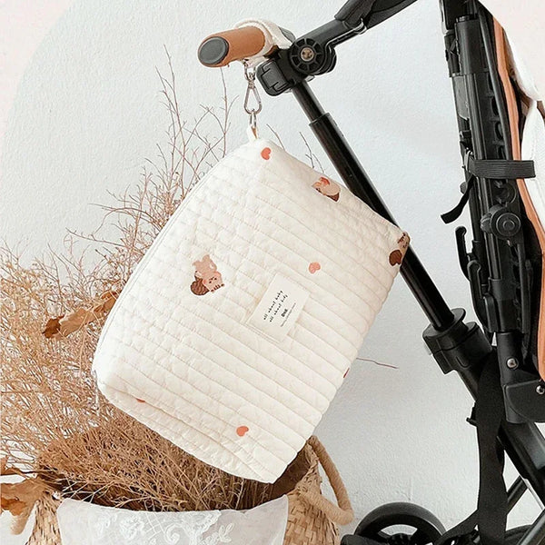 A white quilted cotton diaper and organizer bag is displayed hanging from a black stroller frame.