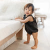A smiling baby wearing a dark denim romper standing next to a white sofa with a fluffy cushion.