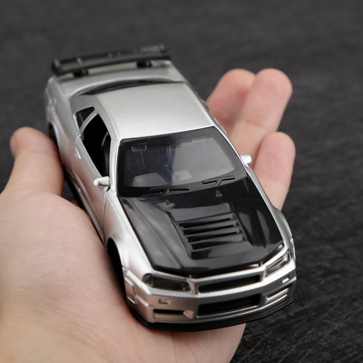 A silver die-cast sports car with a black hood is held in a person's hand.