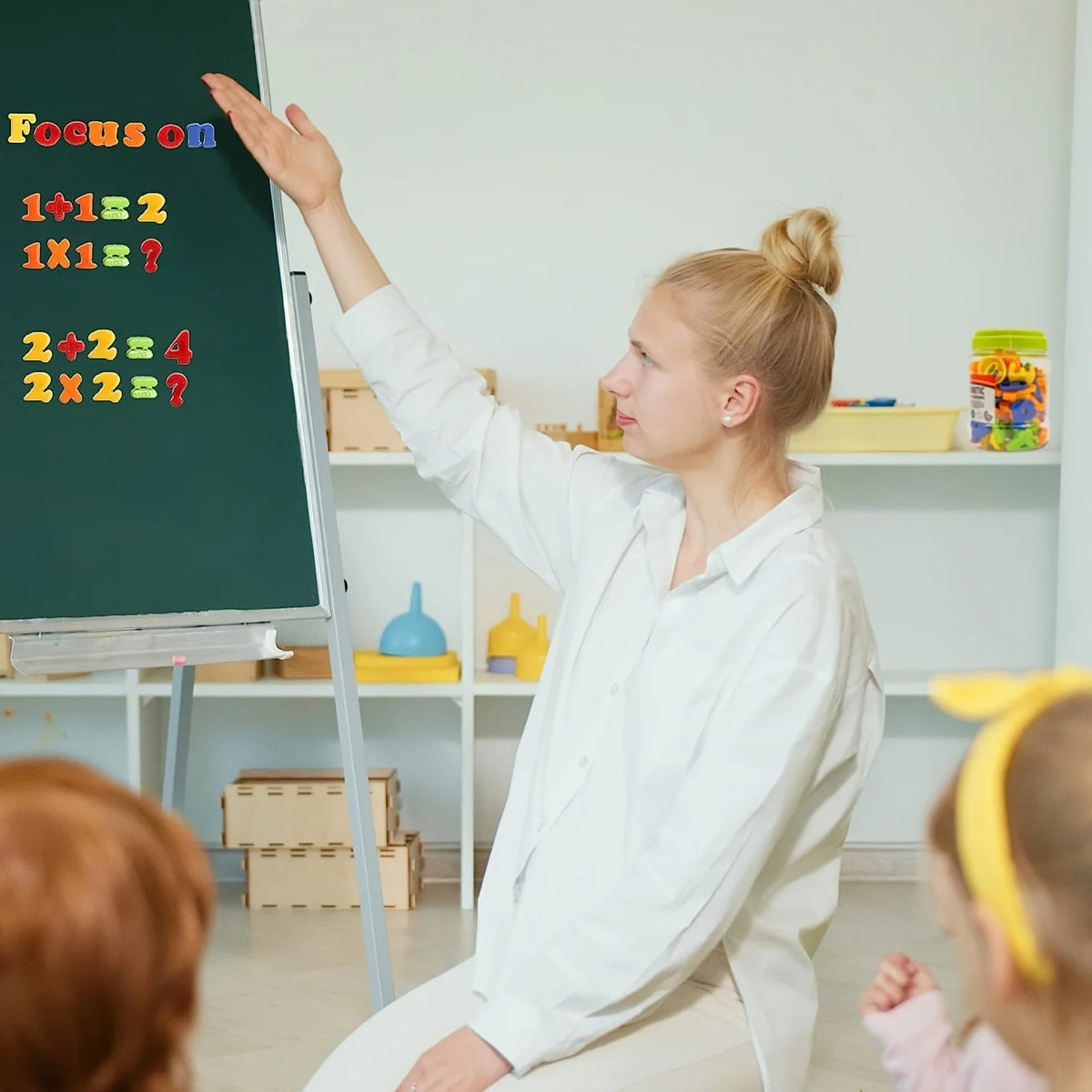 A blonde woman points at a chalkboard with math problems while sitting on the floor in a bright classroom setting.