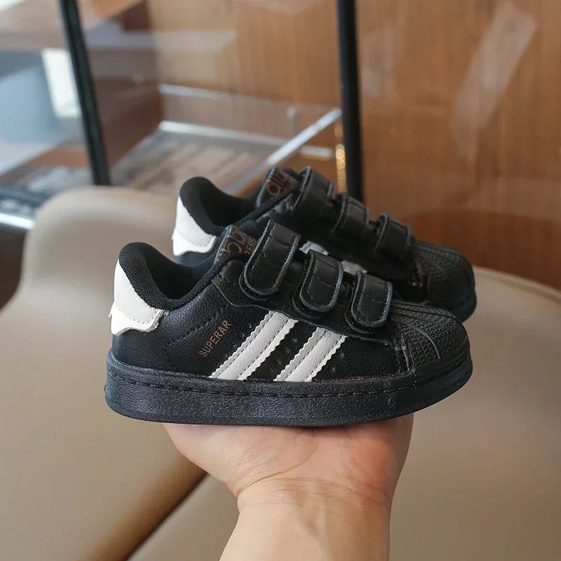 A pair of black and white Adidas sneakers with hook-and-loop closure held in a hand.