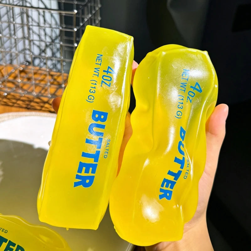 A hand holds a bright yellow butter squishy stress relief toy with the word BUTTER printed on it.