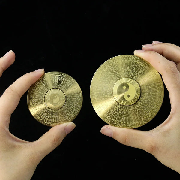A pair of hands holds two round brass Yin Yang compass decor items against a black background.