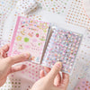A hand holds an open die-cut sticker book featuring a pink cover and a clear sheet of colorful heart stickers.