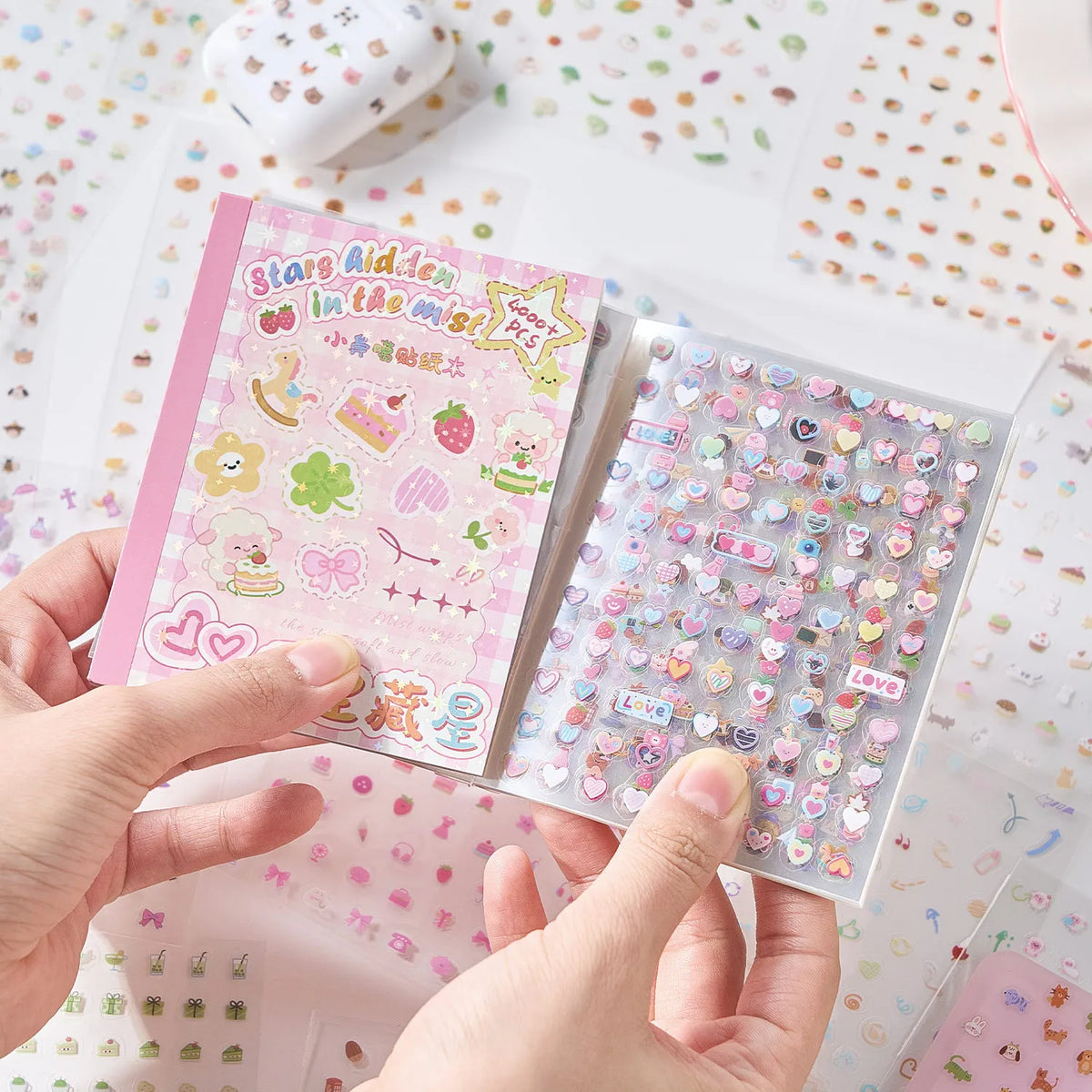A hand holds an open die-cut sticker book featuring a pink cover and a clear sheet of colorful heart stickers.
