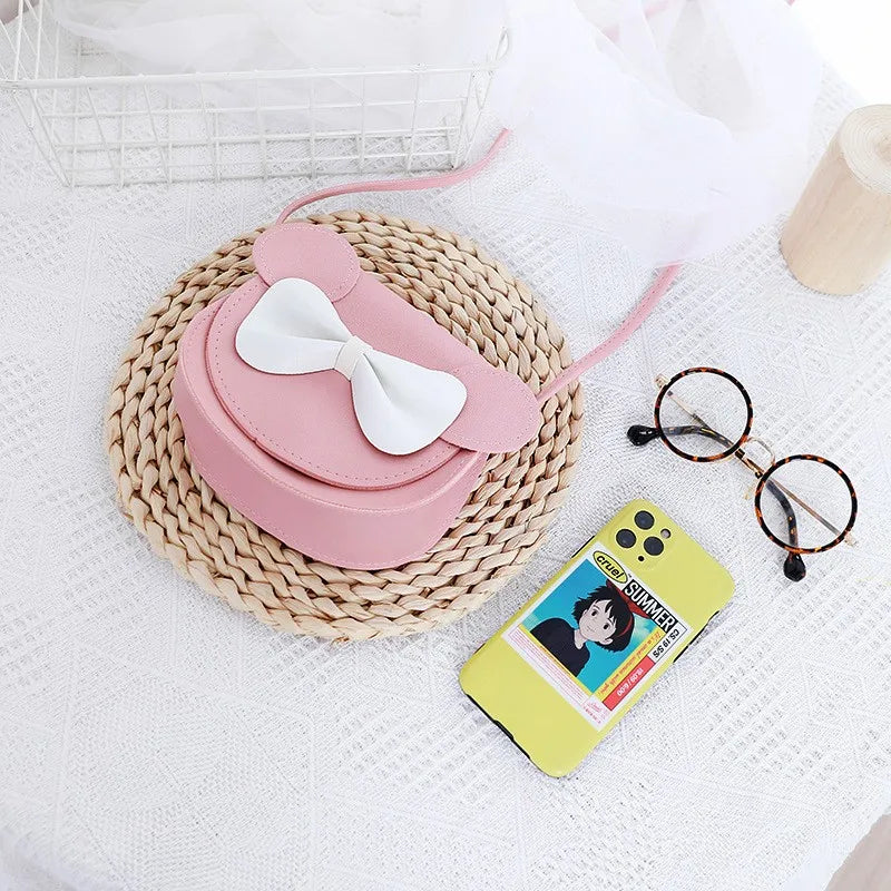 Pink kids hard shell crossbody bag with mouse ears and a white bow resting on a woven mat.