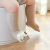 A baby's foot wearing a beige cotton sock with a white cartoon character on a light wooden floor.