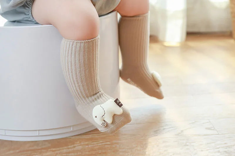 A baby's foot wearing a beige cotton sock with a white cartoon character on a light wooden floor.
