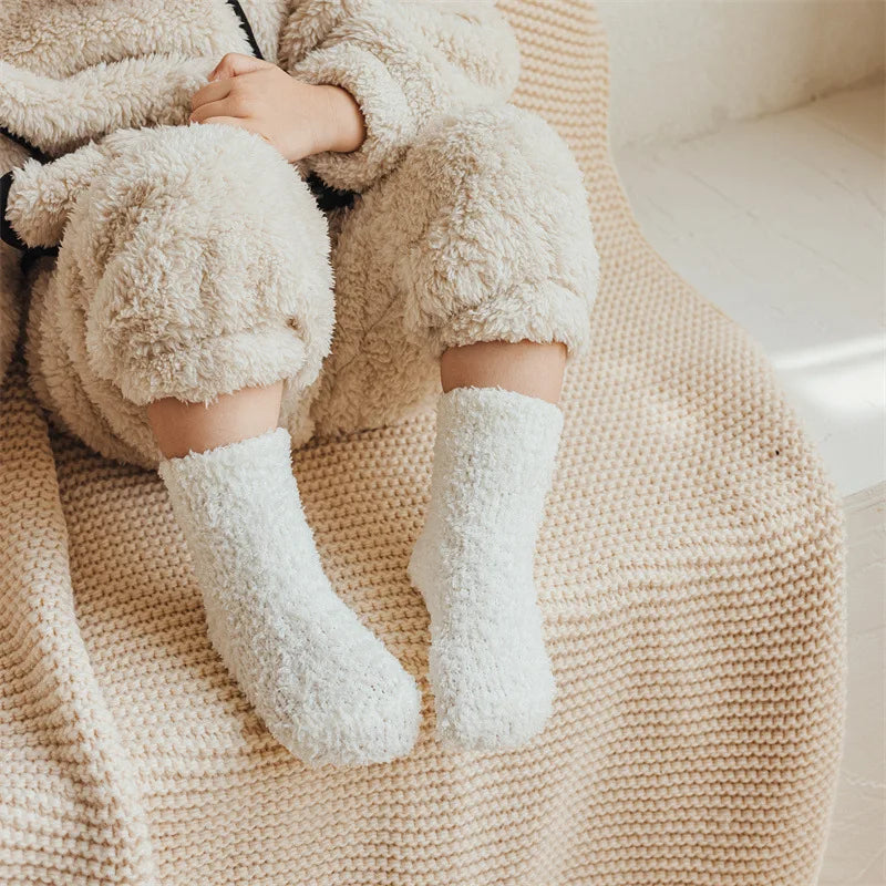 A pair of white fleece socks is shown resting on a beige knitted blanket.