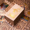 A rectangular box of gold foil tarot cards with a rose pink oracle finish resting on decorative patterned paper.