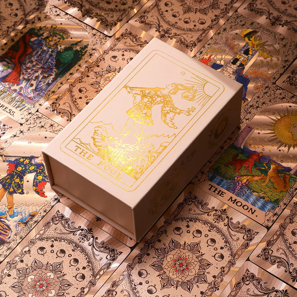 A rectangular box of gold foil tarot cards with a rose pink oracle finish resting on decorative patterned paper.