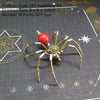 A finished steampunk metal spider assembly kit with eight spindly legs and a red body resting on a dark surface.