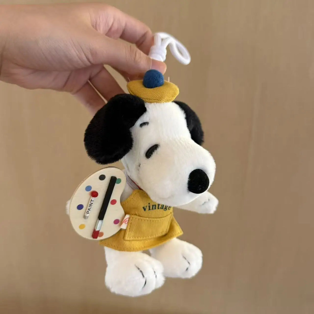 A white Snoopy plush charm with black ears and a yellow vest is being held by a hand.