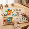 A child interacts with a wooden Montessori busy board featuring gears, switches, and sensory elements on a light wood surface