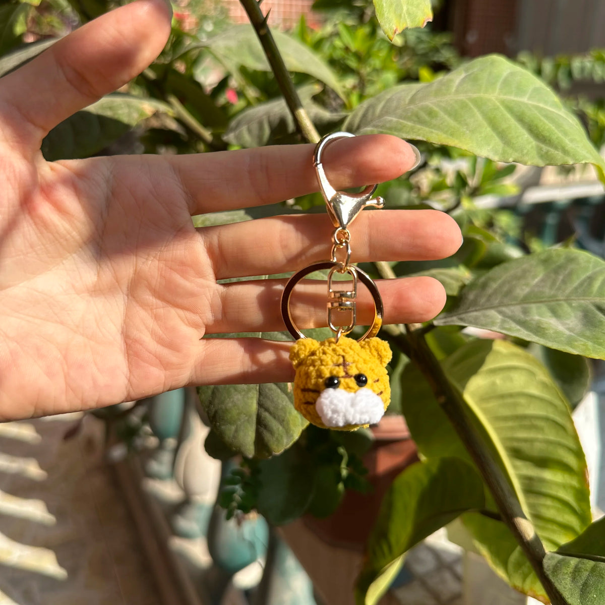 A hand holds a handcrafted crochet animal keychain, resembling a yellow tiger or cat, against a backdrop of green foliage.