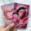 A hand holds a deck of L'Invisible oracle cards featuring a woman wearing a pink mask surrounded by pink peonies and roses.