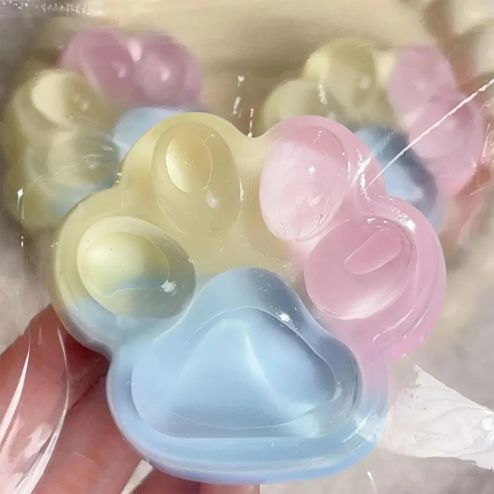 A hand holds a pastel colored squishy toy shaped like a cat paw, featuring light blue and pink translucent silicone.