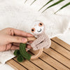 A hand holds a crocheted otter rattle and teether ring with a green leaf detail on a wooden surface.