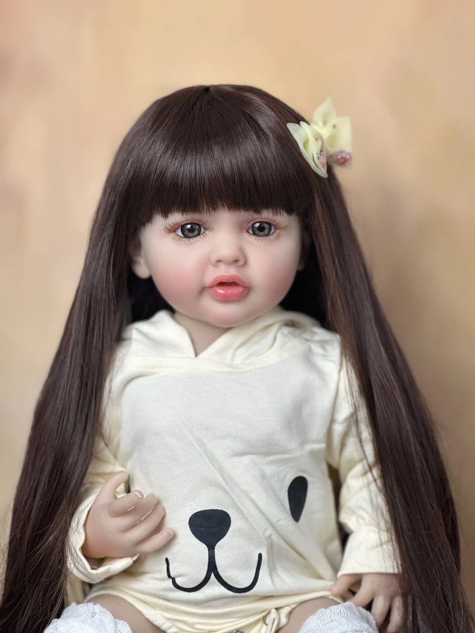 A lifelike 55 cm soft silicone baby doll with long dark hair wearing a cream-colored shirt and a flower accessory.