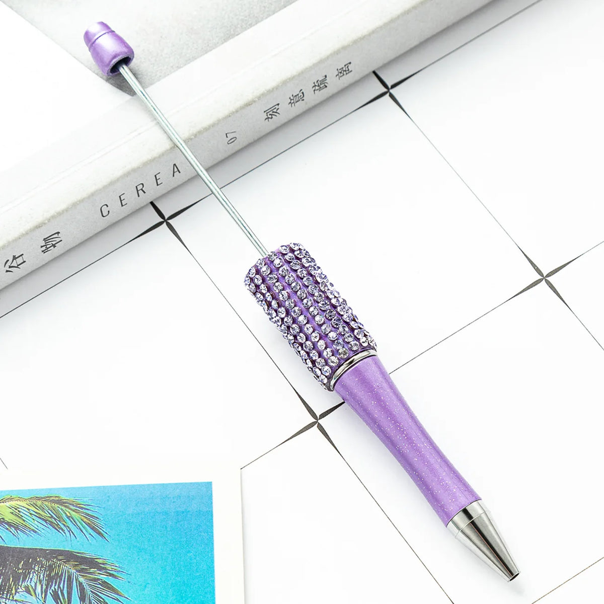 A purple beaded ballpoint pen craft kit with a silver tip rests on a white surface next to a book.