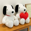 A white plush toy of Snoopy with black ears and a red heart attached, sitting on a wooden surface.