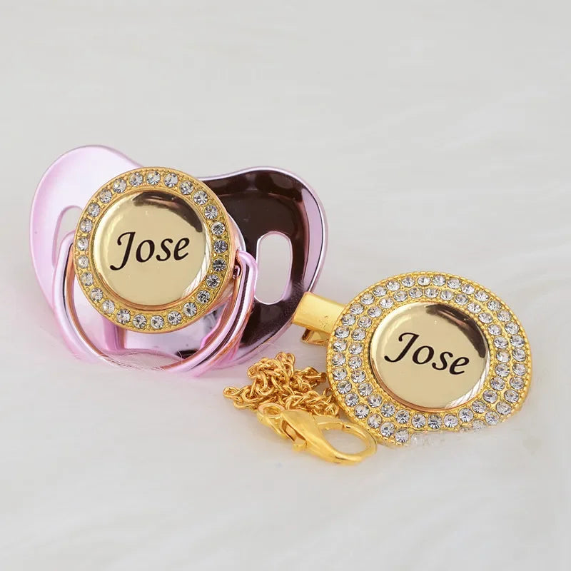 A pink crystal pacifier and clip set with a gold-toned charm featuring the name Jose