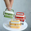 A hand is holding a miniature doll picnic lunch box with a handle, next to a larger red version and some books.
