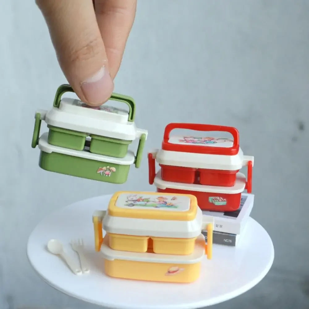 A hand is holding a miniature doll picnic lunch box with a handle, next to a larger red version and some books.