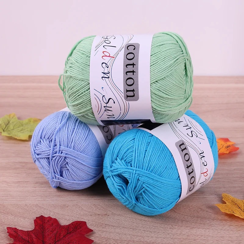 Three skeins of pure cotton knitting yarn in green, blue, and cyan colors are displayed on a wooden surface.