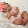 A realistic reborn baby doll with pink clothing and a peach-colored bow lying on a white textured surface.