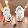 A pair of white soft sole first walker shoes shaped like bears being held by a person.