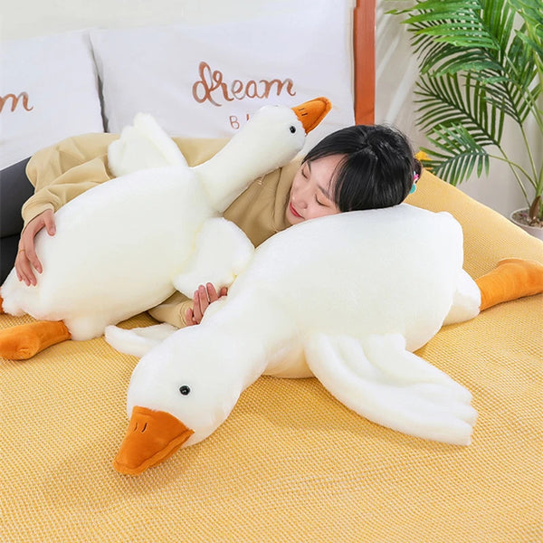 Une personne est allongée avec un grand coussin en peluche de canard blanc, à bec et pattes orange, sur une surface jaune.