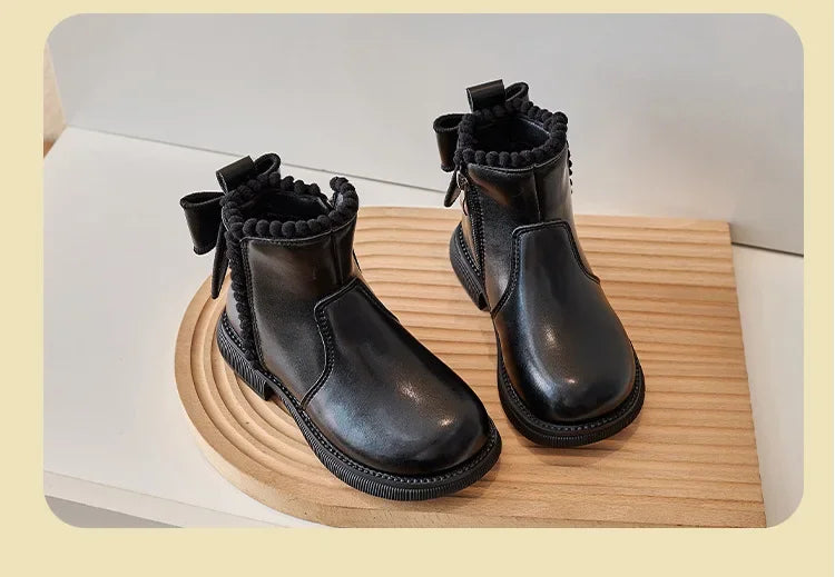 A pair of black girls' ankle boots with a bow-knot detail and side zipper resting on a wooden surface.
