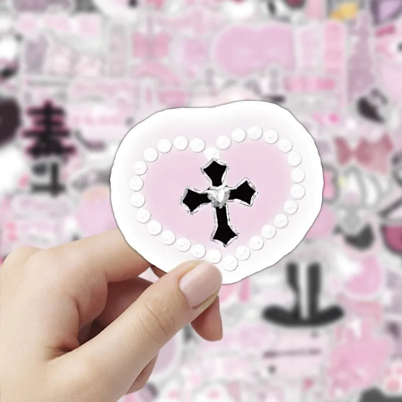 A hand holds a pink heart-shaped sticker with a black cross design on a light background.