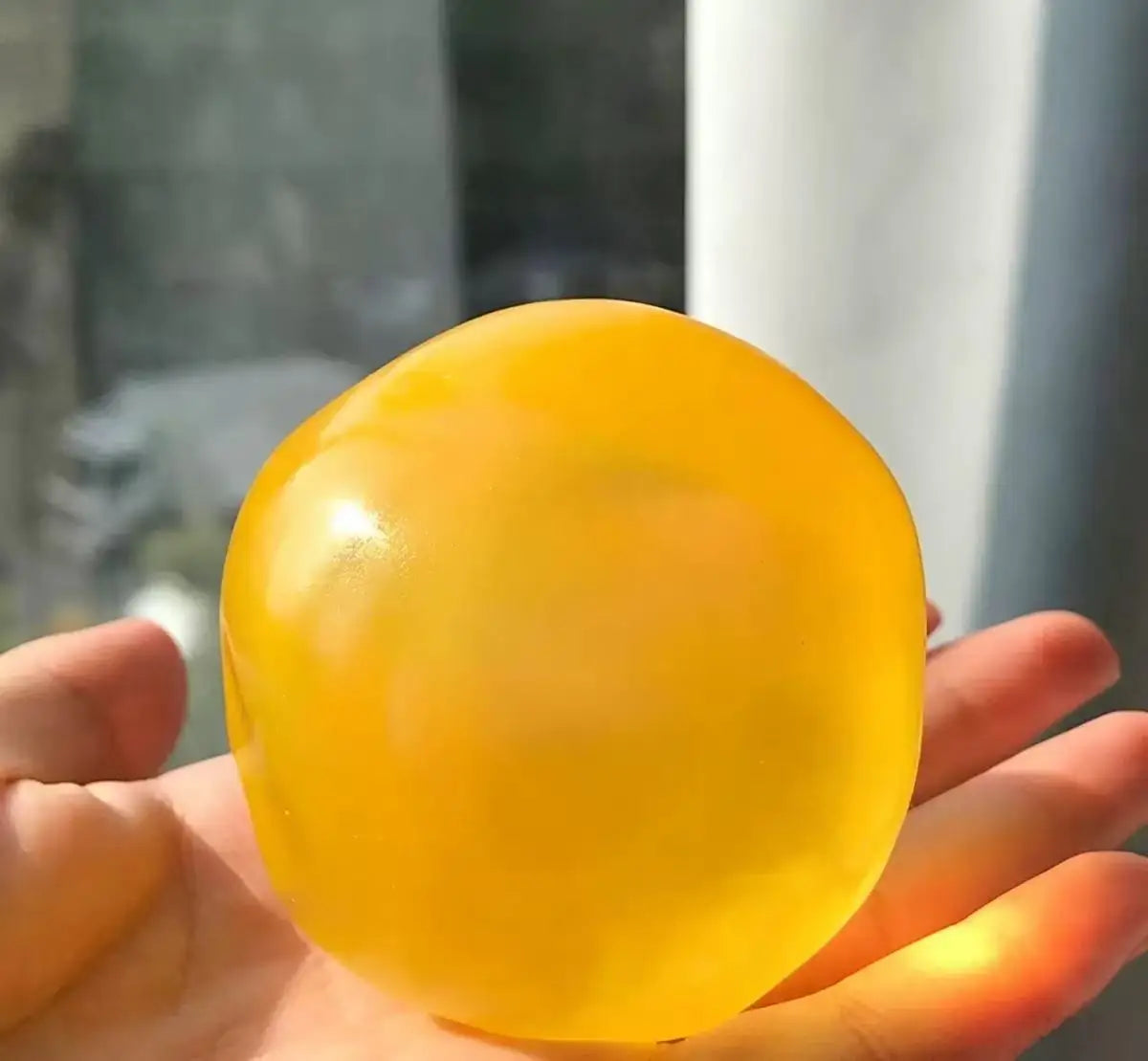 A hand holds a bright yellow, spherical coconut scented slow-rising squishy stress ball.