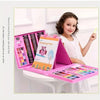 A young girl is using a pink drawing pad and an art set with various colored pencils and crayons on a white table.