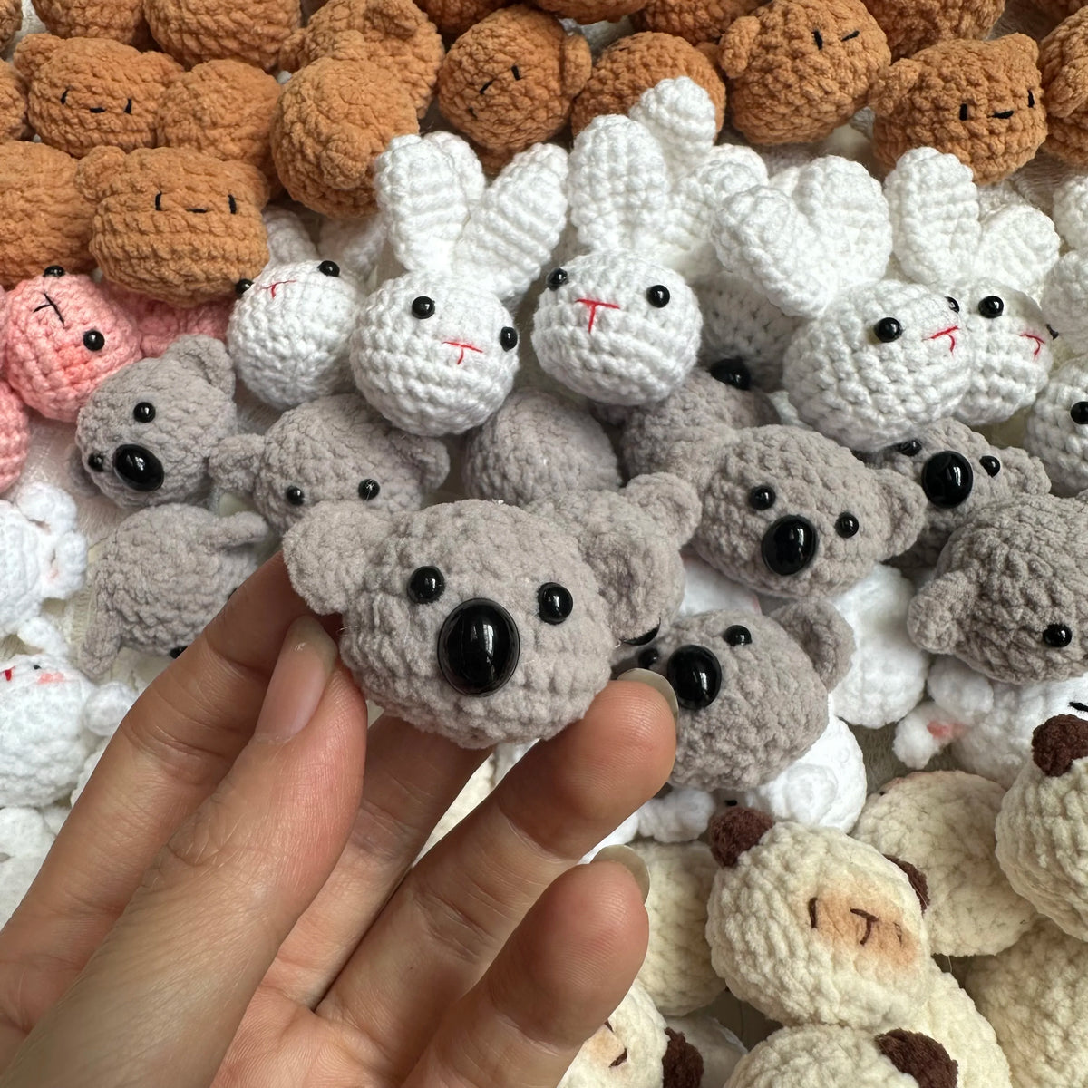 A hand is holding a small, handcrafted crochet animal keychain among many similar soft stuffed animals.