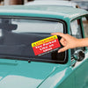 A hand holds up a funny parking violation card with an attention symbol against a teal car.