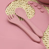 A pink silicone tableware set including a spoon and fork rests on a woven mat.