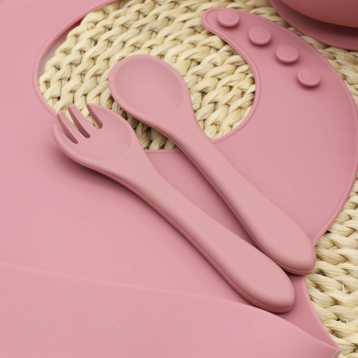 A pink silicone tableware set including a spoon and fork rests on a woven mat.