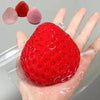 A hand holds a bright red squishy strawberry toy next to several pink and red squishy fruits.