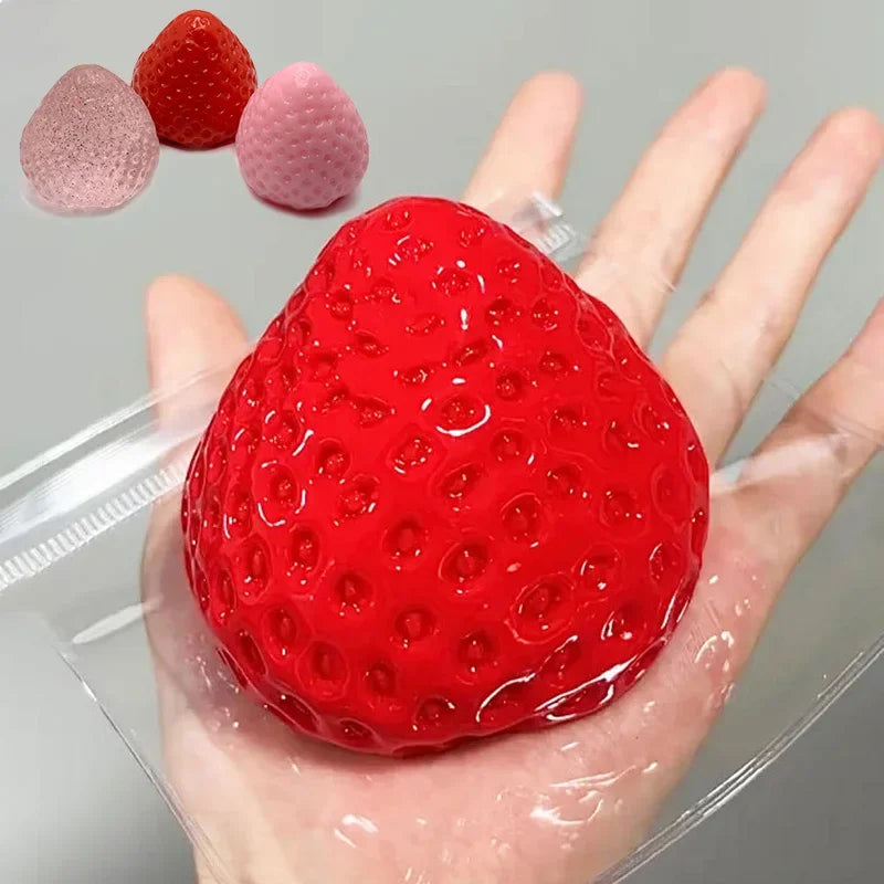 A hand holds a bright red squishy strawberry toy next to several pink and red squishy fruits.