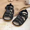 Black and white soft non-slip baby sandals for first walkers resting on a beige fabric.