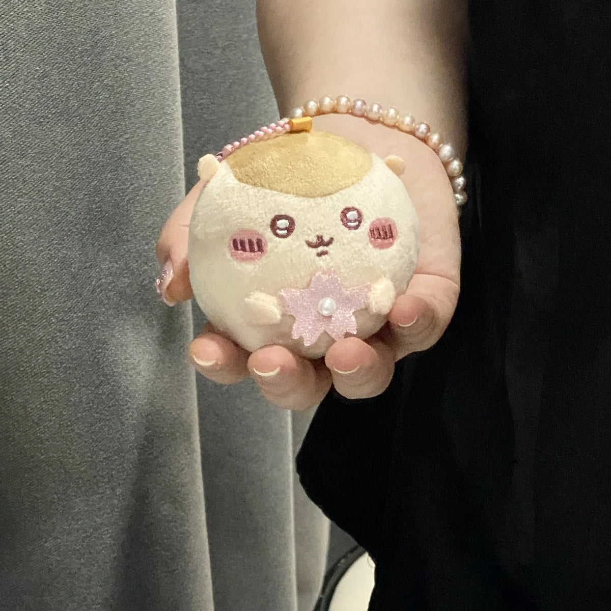 A hand holds a round, light pink plush charm featuring a face and a small pink flower, attached to a beaded bracelet.