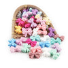 A variety of colorful silicone bow tie beads in pink, purple, yellow, and white are displayed in a wooden tray.