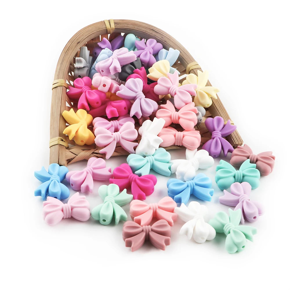 A variety of colorful silicone bow tie beads in pink, purple, yellow, and white are displayed in a wooden tray.