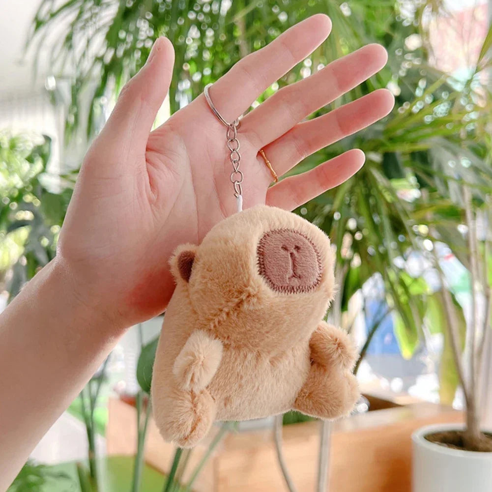 A hand holds a soft, light brown capybara plush keychain charm in a bright, leafy indoor setting.